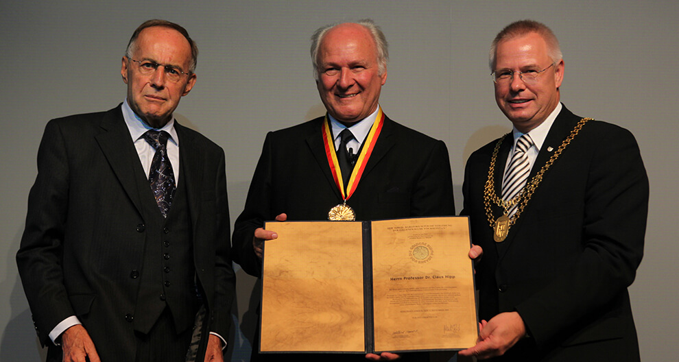 Three men in formal attire stand together, with the central figure holding a large, ornate certificate and a medal.