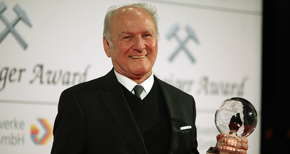 An elderly man in a black suit stands proudly holding a transparent award with a silhouette of a person, against a backdrop that reads “Schweizer Award” and features a logo and a colorful emblem.