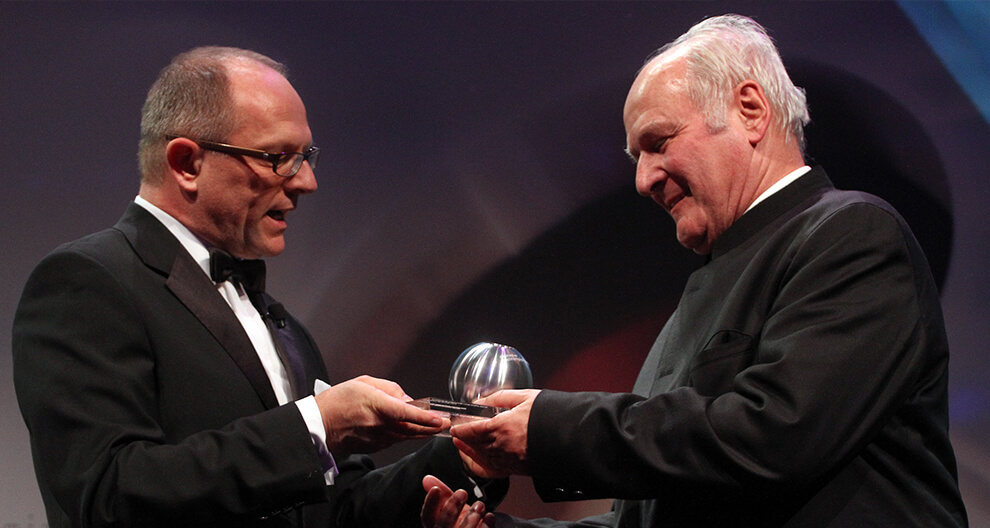 Two men in formal attire exchange a silver spherical award on a stage.