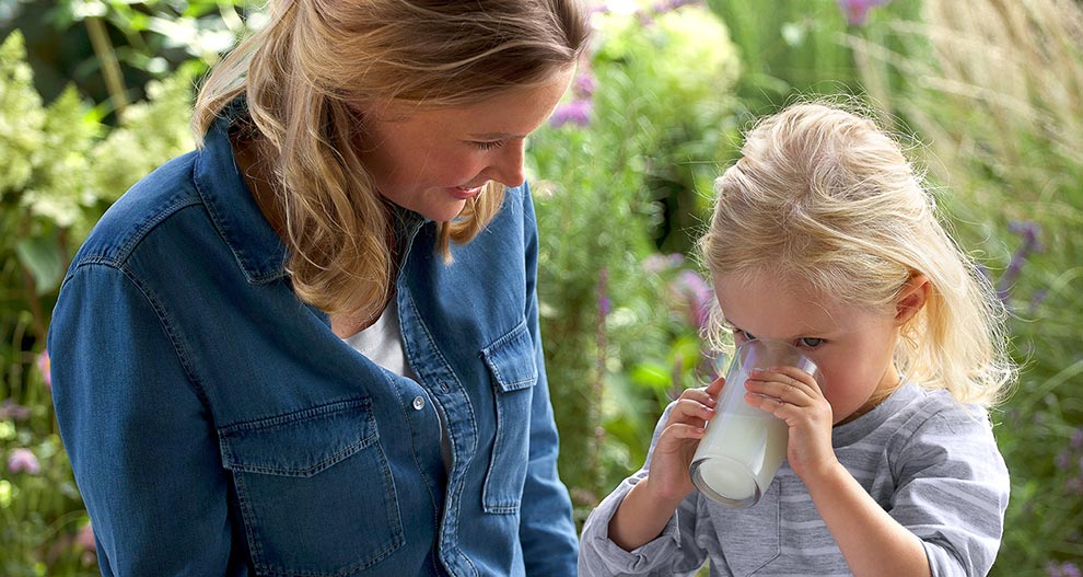 A woman in a blue denim shirt is looking at a young girl drinking from a glass.