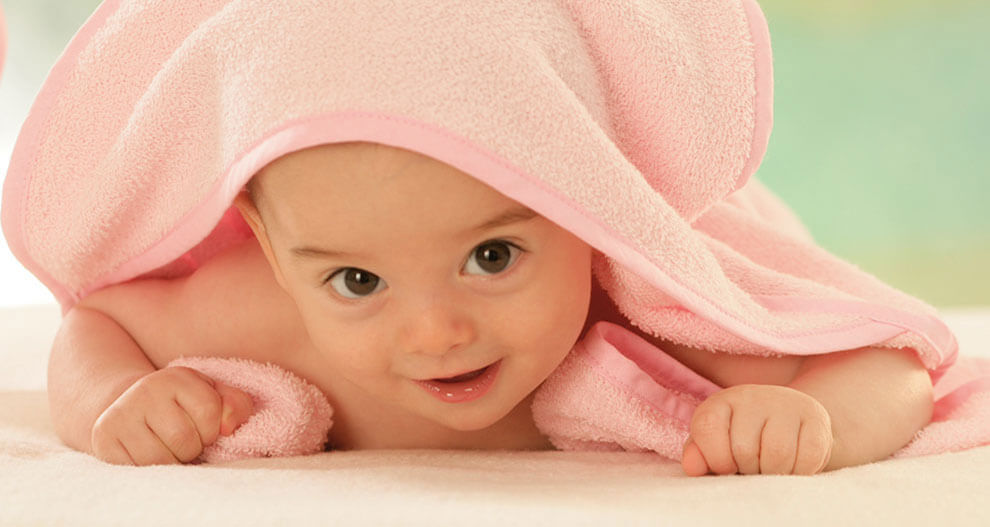 A baby wrapped in a pink towel lies on a white surface, looking directly at the camera.