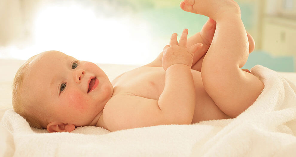 A baby lies on a white towel, looking up with a hand raised.