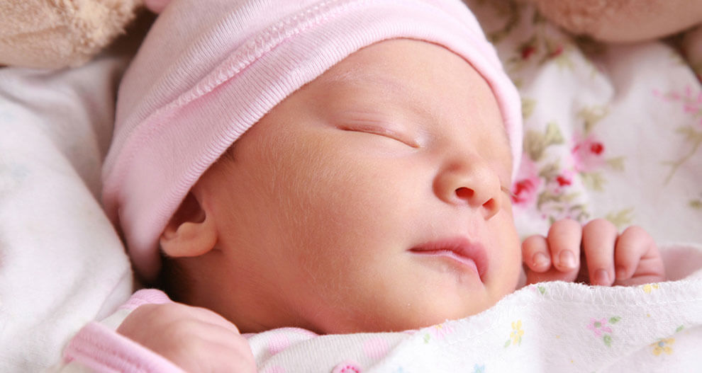 A close-up of a sleeping baby's face, wearing a pink hat, surrounded by soft fabric with floral patterns.