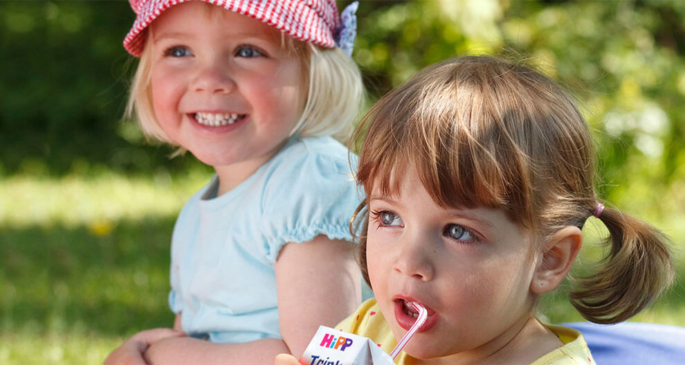Two young girls sit outdoors, one wearing a red and white checkered hat, the other sipping from a white carton with a straw.