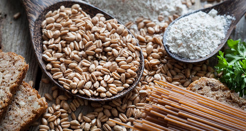 A wooden table displays a variety of whole grain foods, including uncooked spaghetti, a pile of wheat grains, a slice of rustic bread, and a heap of flour.