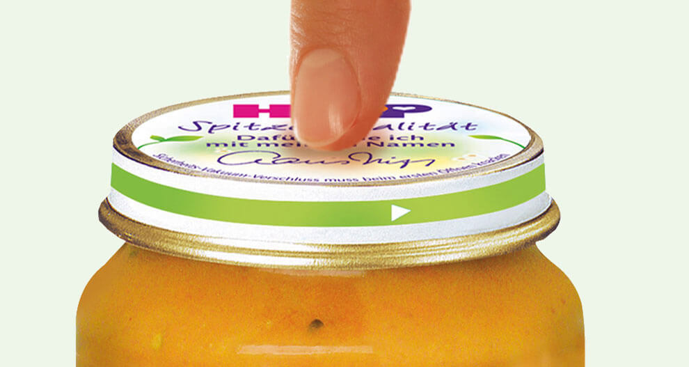 A hand is about to open a jar of yellow food product with a green lid.