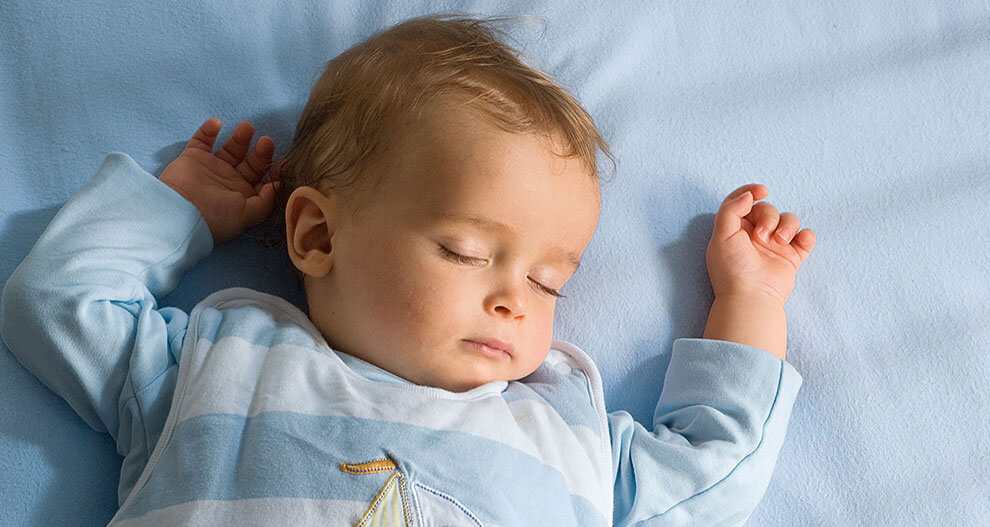 A baby lies on a blue surface, wearing a light blue and white striped outfit with a sailboat design.