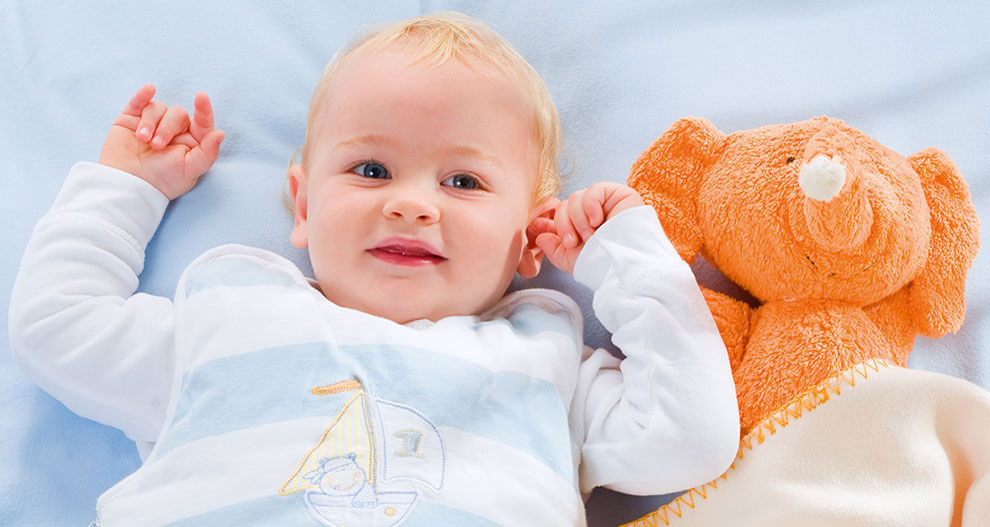 A baby lies on a blue blanket, holding a plush elephant toy.