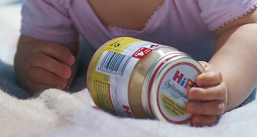 A baby's hand holds a jar with a barcode and label.
