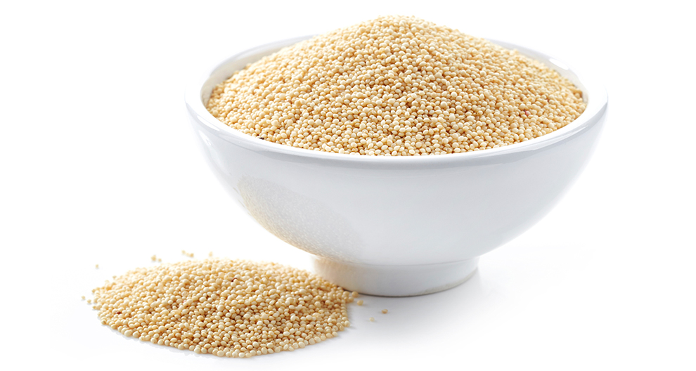 A white bowl filled with small, round, beige grains is placed on a white surface, with a smaller pile of the same grains beside it.
