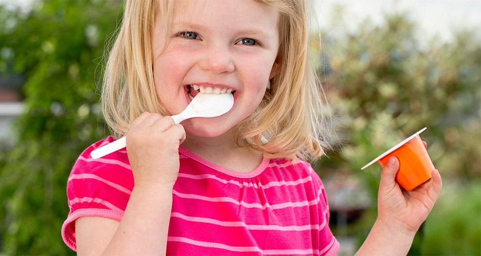 A girl in a pink-striped shirt is eating from an orange container with a white spoon.
