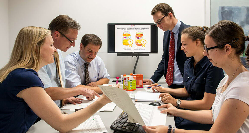 A group of professionals is engaged in a meeting, with a focus on a presentation displayed on a computer screen that features a product advertisement.