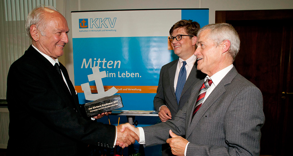 Three men in suits are in a room with a blue banner displaying the letters "KKV" and German text, with one man receiving an award from another.