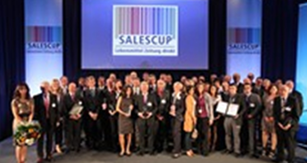 A group of individuals stands on a stage, holding awards and certificates, with a large screen displaying the word "SALESCUP" in the background.