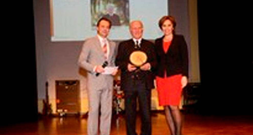 Three individuals stand on a stage, with one holding a round, wooden award.