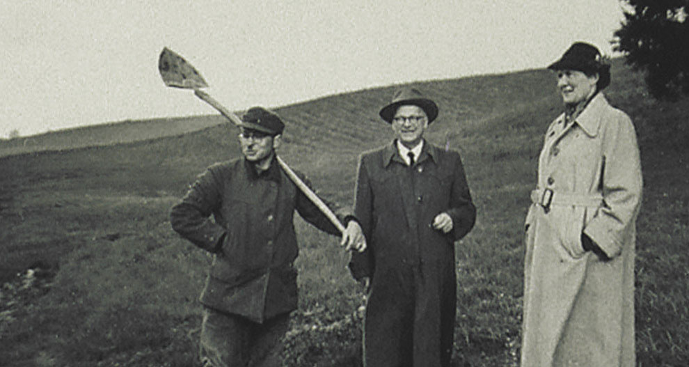 Three individuals stand in an open field; one holds a shovel, and the others are dressed in formal attire.